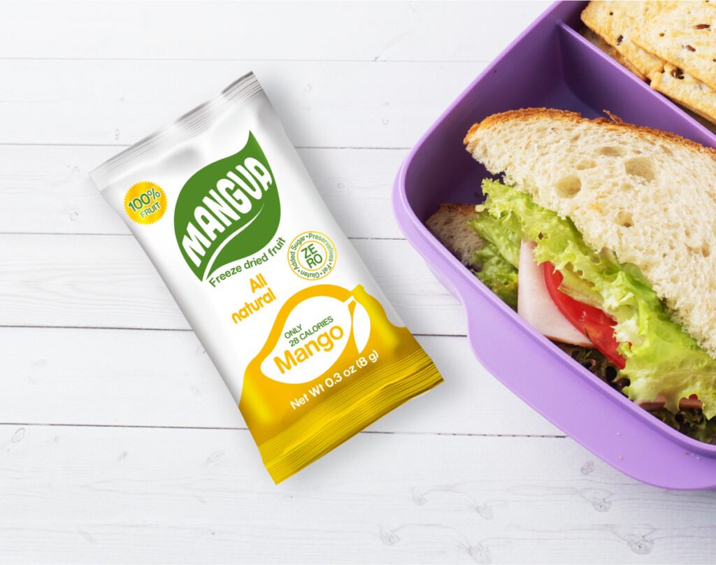 MANGUA freeze-dried mango snack on a white wooden table beside a purple lunchbox with a sandwich, ideal for healthy, natural snacking without added sugar.